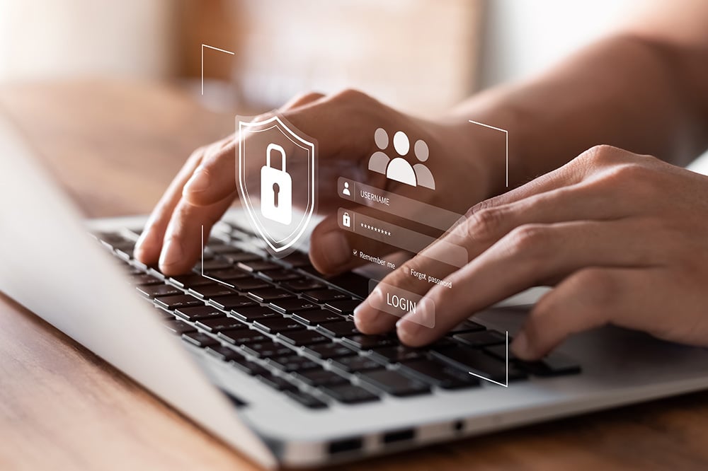 AdobeStock_460630045 Close-up of hands typing on a laptop keyboard with digital graphics of a lock, shield, and login fields, symbolizing cybersecurity and secure online access.